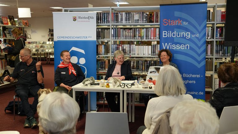 Ein Tisch an dem drei Damen sitzen. Im Hintergrund ein Büchereiregal und im Vordergrund ältere Menschen, die einer der Damen am Tisch beim Vorlesen zuhören.