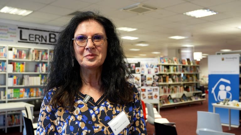 Eine Frau mittleren Alters, mit langen, schwarzen, lockigen Haaren. Trägt ein buntes Shirt und steht in einer Bibliothek. 