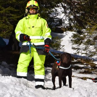 Frau trägt einen leuchtgelben Anzug mit Reflektoren und einen gelben Helm. Mit der linken Hand hält sie ihren dunkelbraunen Hund an der Leine fest. Beide stehen im Schnee. 