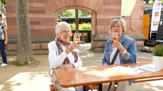 Zwei ältere Damen sitzen an einem Tisch im Freien und halten Weingläser hoch.