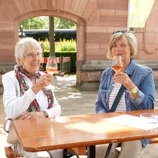 Zwei ältere Damen sitzen an einem Tisch im Freien und halten Weingläser hoch.