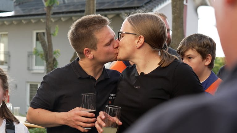 Junge Frau küsst einen jungen Mann mit kurzen Haaren. Beide tragen ein schwarzes T-Shirt.