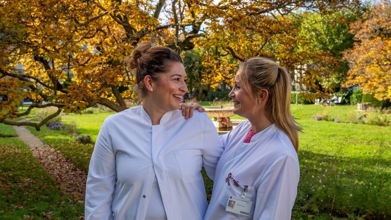 Zwei Frauen in weißen Kitteln lächeln sich an und stehen in einem Park.