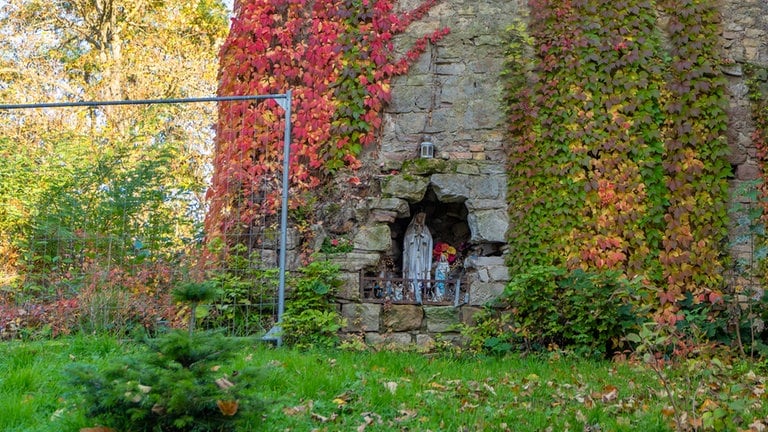 In einer alten Mauer ist in einer Nische ein Schrein aufgebaut. Bunter Efeu rankt sich an der Mauer empor und ein Bauzaun ragt links ins Bild.