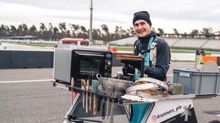 Junger man beim Marathonlaufen in einem Gestellt mit Rädern, auf dem ereinen Kuchen backt.