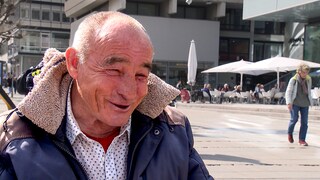 Rudolf spielt jeden Tag Schach auf dem Schlossplatz in Stuttgart. Er sitzt an seinem Klapptisch mit seinem Schachbrett und lacht in die Kamera.