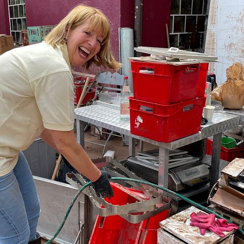 Dorothe und ihre Metzgerei in Rudersberg sind vom Hochwasser betroffen. Dorothe räumt die Schäden auf und greift nach einer roten Kiste. Sie lacht in die Kamera. 