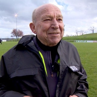 Umberto ist seit über 50 Jahren Schiedsrichter im Amateur-Fußball. Er steht in seiner Schiedsrichter-Ausrüstung auf einem Fußballfeld und lacht in die Kamera.