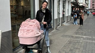 Maria steht in der Fußgängerzone ganz in der Nähe des Stuttgarter Rathauses und schiebt ihren rosa Kinderwagen, der mit vielen Blumen verziert ist. Maria lächelt in die Kamera. 