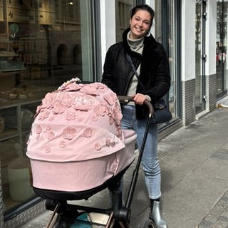 Maria steht in der Fußgängerzone ganz in der Nähe des Stuttgarter Rathauses und schiebt ihren rosa Kinderwagen, der mit vielen Blumen verziert ist. Maria lächelt in die Kamera. 