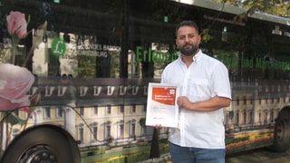 Busfahrer Mustafa steht vor seinem Bus und zeigt stolz seinen Preis für Zivilcourage in die Kamera.