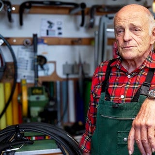 Der 79-Jährige Roland steht in seiner Fahrradwerkstatt. An den Wänden hängen historische Fahrradrahmen und Ersatzteile und erzählt von seiner Arbeit.