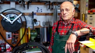 Der 79-Jährige Roland steht in seiner Fahrradwerkstatt. An den Wänden hängen historische Fahrradrahmen und Ersatzteile und erzählt von seiner Arbeit.