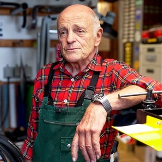 Der 79-Jährige Roland steht in seiner Fahrradwerkstatt. An den Wänden hängen historische Fahrradrahmen und Ersatzteile und erzählt von seiner Arbeit.