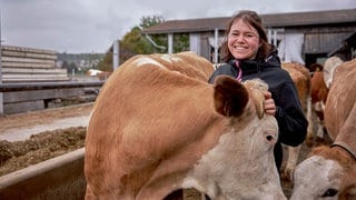 Landwirtin Monja steht mit einer ihrer Kühen im Außenbereich des Kuhstalls. Sie lächelt in die Kamera und hält die Kuh am Kopf.