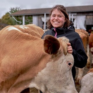 Landwirtin Monja steht mit einer ihrer Kühen im Außenbereich des Kuhstalls. Sie lächelt in die Kamera und hält die Kuh am Kopf.