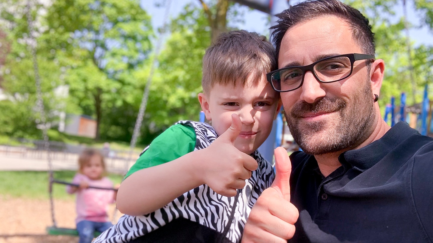 Tagesvater Saky und sein Sohn sind auf dem Spielplatz. Sie machen ein Selfie und lachen mit Daumen nach oben in die Kamera.