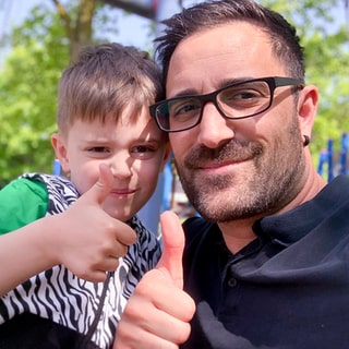 Tagesvater Saky und sein Sohn sind auf dem Spielplatz. Sie machen ein Selfie und lachen mit Daumen nach oben in die Kamera.