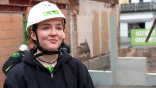 Martha ist 18 Jahre alt und macht nach ihrem Abitur eine Ausbildung zur Stahlbetonbauerin. Sie steht im Vordergrund und schaut in die Kamera. Sie trägt einen Helm und im Hintergrund sind ihre Kollegen zu sehen.