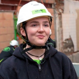 Martha ist 18 Jahre alt und macht nach ihrem Abitur eine Ausbildung zur Stahlbetonbauerin. Sie steht im Vordergrund und schaut in die Kamera. Sie trägt einen Helm und im Hintergrund sind ihre Kollegen zu sehen.