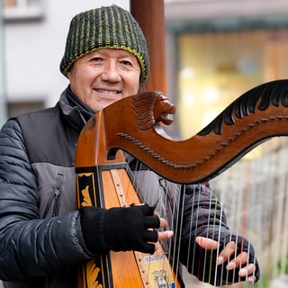 Harfenist Milton steht in der Esslinger Innenstadt. Er hält seine Harfe fest und lacht in die Kamera.