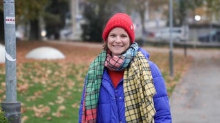 Mama Lea, nachdem sie auf einem Spielplatz mit ihren zwei Kindern gespielt hat. Sie lächelt in die Kamera, trägt eine rote Mütze, einen karierten Schal und eine blaue Jacke.