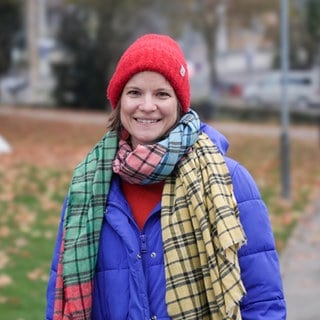 Mama Lea, nachdem sie auf einem Spielplatz mit ihren zwei Kindern gespielt hat. Sie lächelt in die Kamera, trägt eine rote Mütze, einen karierten Schal und eine blaue Jacke.