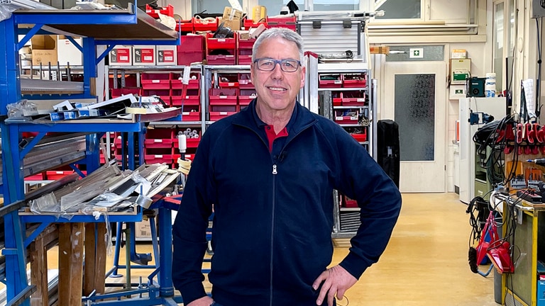 Uwe arbeitet seit 50 Jahren im selben Betrieb. Er steht in der Werkstatt eines Fensterbau-Unternehmens. Er lächelt in die Kamera. 