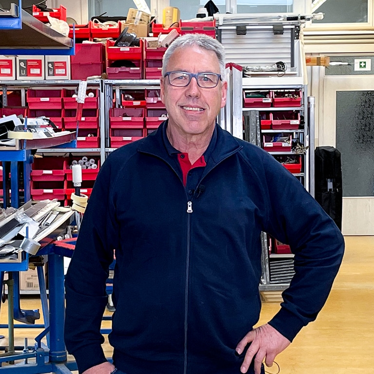 Uwe arbeitet seit 50 Jahren im selben Betrieb. Er steht in der Werkstatt eines Fensterbau-Unternehmens. Er lächelt in die Kamera. 