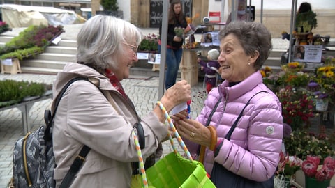 Die 78-jährige Carin verschenkt einen Glücksbringer an eine Seniorin auf dem Markt. Beide lachen herzlich.