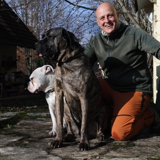 Hundetrainer Michael setzt sich für die Rehabilitation von Hunden ein. Er kniet auf den Boden und hält drei Hunde an ihren Halsbändern fest. Er lacht in die Kamera.