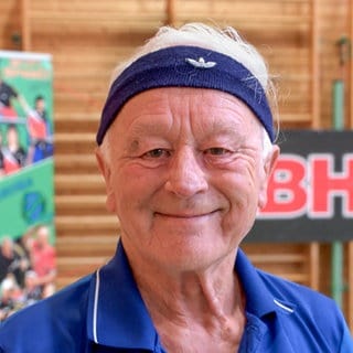 Hans ist bei einem Tischtennistraining und lächelt in die Kamera. Er trägt ein blaues Stirnband und Sportkleidung. 