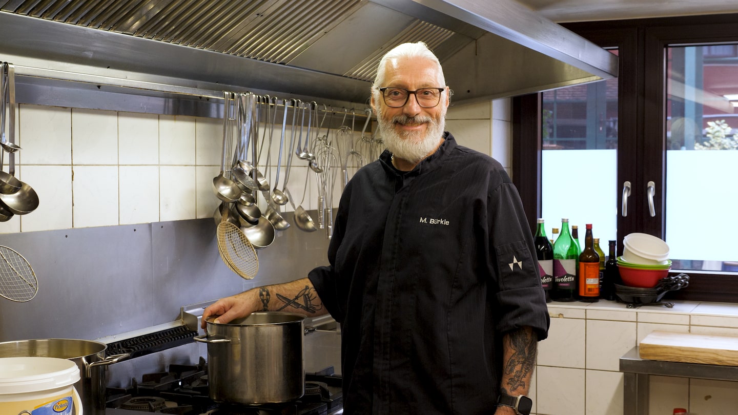 Restaurantbesitzer Marc kocht ehrenamtlich für Sterbende im Hospiz. Er steht in der Küche am Herd in seinem Restaurant. Auf dem Herd steht ein Topf. Er trägt schwarze Sachen und lacht in die Kamera.