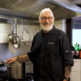 Restaurantbesitzer Marc kocht ehrenamtlich für Sterbende im Hospiz. Er steht in der Küche am Herd in seinem Restaurant. Auf dem Herd steht ein Topf. Er trägt schwarze Sachen und lacht in die Kamera.