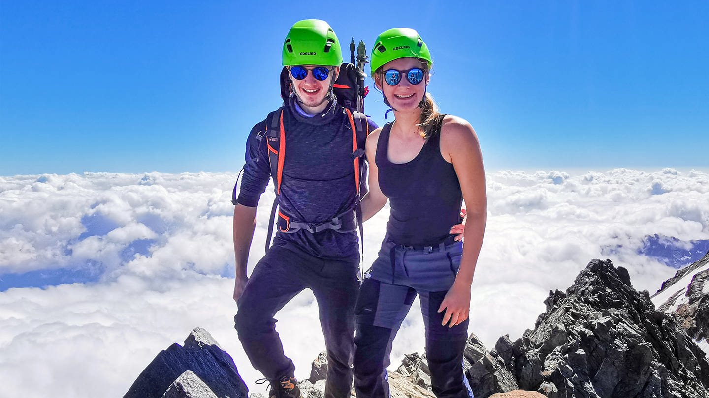 Zwanzigjähirge/r Frau und Mann stehen mit Wanderausrüstung auf dem Gipfel eines Berges.