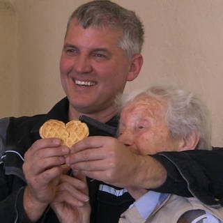 Ein Mann und eine Seniorin halten ein Waffel-Herz in der Hand und lächeln