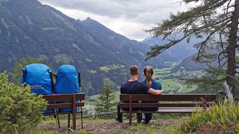 Zwanzigjähriges Pärchen sitzt Arm in Arm auf einer Bank und schaut in die Ferne. Auf der Bank daneben stehen  Wanderrucksäcke und  Wanderstöcke. 