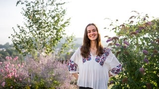 Stella in weiß-geblümten Shirt vor einer grünen Hecke