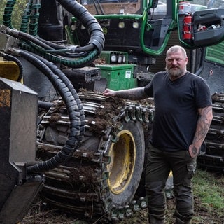 Ein Mann steht vor einem großen Harvester und hält seine Hand auf der Kette