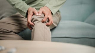 Ein Mann sitzt auf einem grauen Sofa und fasst sich ans Knie. Er trägt ein grünes Hemd und eine beige Hose. 