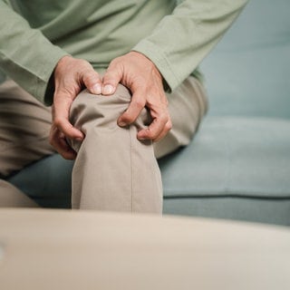 Ein Mann sitzt auf einem grauen Sofa und fasst sich ans Knie. Er trägt ein grünes Hemd und eine beige Hose. 