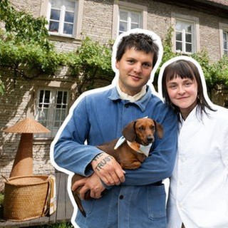 Pat und Sissi sind ein junges Paar und stehen mit einem Dackel im Arm vor einem alten Haus in Unterfranken.