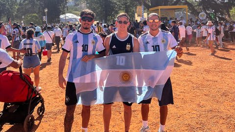 Dominic Sessa (r.) steht mit seinen Brüdern Kevin (l.) und Nicolas (m.) auf einem Public Viewing Feld und halten die argentinische Flagge in die Kamera.