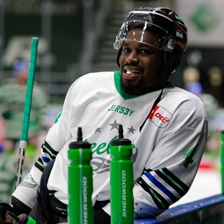 Der Schwarze Eishockeyspieler Jalen Smereck lehnt an der Bande der Ersatzbank der Bietigheim Steelers und schaut auf die vor ihm aufgereihten grünen Getränkeflaschen.