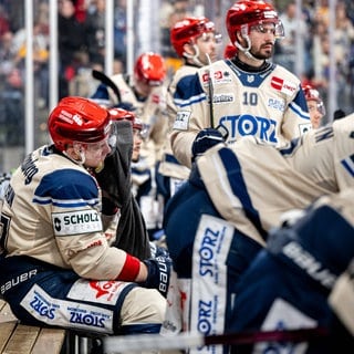 Die Schwenninger Eishockeyspieler sitzen auf der Auswechselbank und beobachten das Spiel.