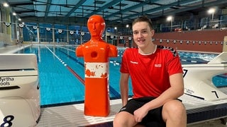 Leo Baumann sitzt am Beckenrand zwischen zwei Startblöcken. Links von ihm steht eine rot-weiße Puppe, die er zuvor aus dem Wasser "gerettet" hat. Im Hintergrund ist das Schwimmbecken zu sehen.