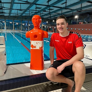 Leo Baumann sitzt am Beckenrand zwischen zwei Startblöcken. Links von ihm steht eine rot-weiße Puppe, die er zuvor aus dem Wasser "gerettet" hat. Im Hintergrund ist das Schwimmbecken zu sehen.