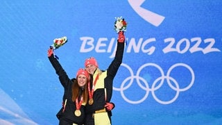 Katharina Hennig (l.) und Victoria Carl (r.) stehen bei der Siegerehrung vor einer blauen Wand, auf der Beijng 2022 steht. Um den Hals tragen sie Goldmedaillen und sie recken Blumensträuße in die Luft.