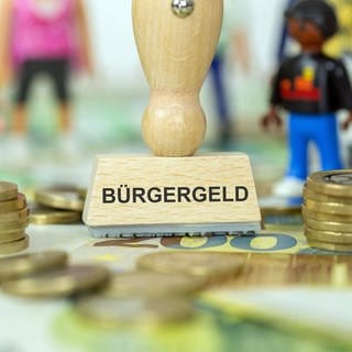 Ein Symbolbild für Bürgergeld - mit einem Stempel mit der Aufschrift "Bürgergeld", mit Playmobil Spielzeugfiguren und Eurobanknoten