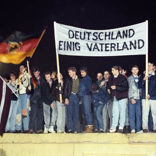 Mit der Deutschlandfahne und einem Transparent "Deutschland Einig Vaterland" stehen zahlreiche Berliner am 22.12.1989 auf der Berliner Mauer am Brandenburger Tor.